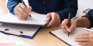 Two people at wooden desk writing down things on clipboard and notebook